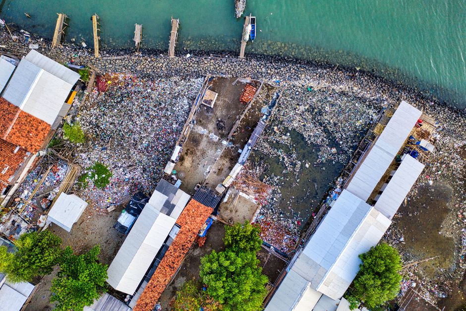 An aerial view of a coastal area showing a large accumulation of waste and debris scattered across a narrow shoreline, with trash spilling from nearby structures and onto the ground. The waste consists of various materials including plastic bottles, packaging, and other refuse, creating a dense, mixed layer along the edge of the water. Several buildings with white and red-tiled roofs are positioned adjacent to the contaminated area, and some structures appear to have overhanging roofs or scaffolding. The rocky shoreline is lined with a concrete or stone barrier, separating the waste from the water, which features a few small boats docked near the edge. Vibrant green trees are visible in the lower part of the image, contrasting with the cluttered, rubbish-strewn environment. The overall scene depicts a significant pollution problem impacting the coastline, illustrating the importance of proper waste disposal, such as private rubbish clearance services provided by companies like Rubbish Clearance Kingston, especially in scenarios where independent collection or on-site clearance may be necessary.