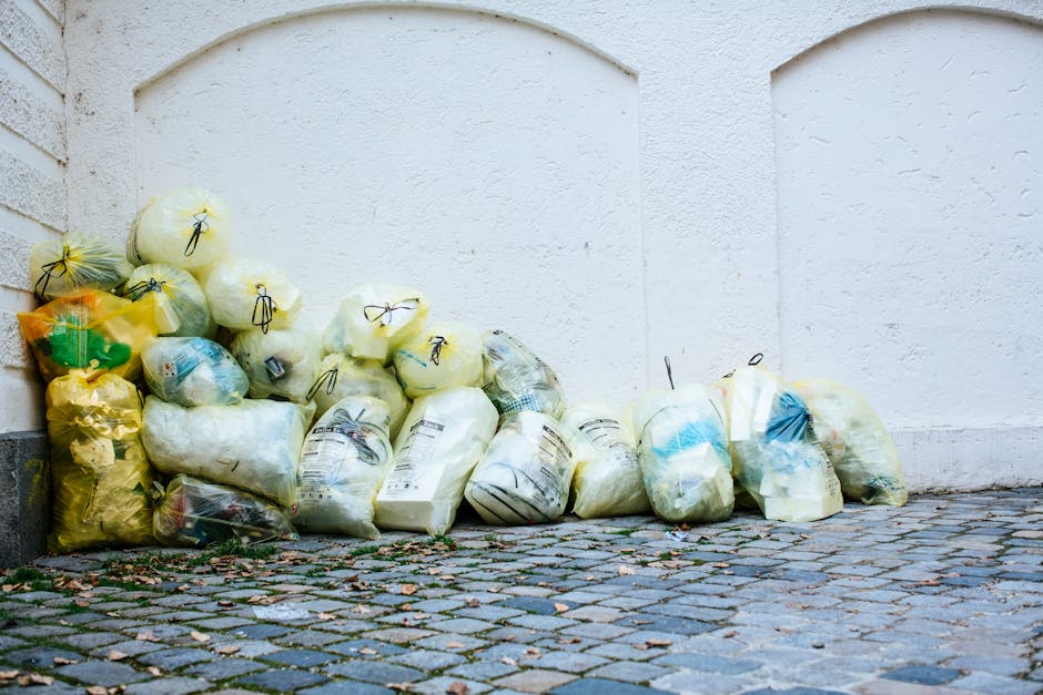A row of tightly gathered plastic rubbish bags, predominantly yellow, white, and clear, are piled against a white, textured exterior wall near the corner of a brick building. The bags contain miscellaneous waste materials, with some visibly bulging and tied with black or yellow ties. The bags are placed on a cobblestone pavement with small, uneven stones, and a few fallen leaves scattered around. The background features a plain white wall with subtle surface imperfections, and the scene appears to be outdoors, illuminated by natural light. The image depicts a typical scenario of waste awaiting collection or disposal, illustrating the kind of rubbish that might be handled by independent waste removal services such as those provided by Rubbish Clearance Kingston, especially in the context of private waste management or on-site clearance for properties in Norbiton.