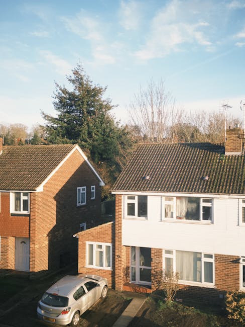 A view of two adjacent residential houses situated in a suburban neighbourhood under a clear blue sky with some light clouds. The house on the left features a brown brick exterior with white-framed windows and a tiled roof with a slight overhang. The house on the right has a combination of brick on the ground floor and white cladding on the upper floor, also with a tiled roof. In front of the houses, a driveway is visible with a silver car parked towards the lower left corner. The foreground shows a small garden with a young shrub and a concrete pathway leading to the entrance doors. Behind the houses, there are tall trees with bare branches and more distant trees with sparse leaves, suggesting the season is late autumn or early winter. The scene is illuminated by natural daylight, highlighting the textures of the brickwork, roofing tiles, and foliage, consistent with a typical property scene that could involve private waste collection or on-site clearance activities by rubbish removal services such as Rubbish Clearance Kingston.