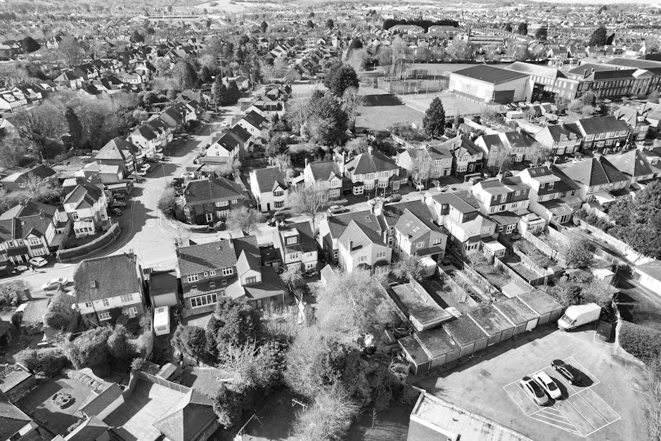An aerial black-and-white photograph of a residential suburban area showing a dense arrangement of semi-detached and terraced houses with pitched roofs, some with solar panels and chimneys. The streets are narrow and winding, lined with parked cars and small front gardens with trees and shrubs. In the foreground, there is a yard with multiple garden sheds, fence panels, and various cluttered debris, indicating a cluttered outdoor space awaiting clearance. Surrounding the yards are trees and hedges, some casting shadows. Beyond the residential units, a larger sports field with a fence and sports equipment is visible, along with a school or community hall building nearby. In the background, the expansive townscape stretches towards the horizon, with densely packed houses, roads, and scattered green spaces. The scene suggests a typical urban neighborhood where separate rubbish or waste collection services, such as those offered by Rubbish Clearance Kingston, could assist in private waste disposal or yard clearance, aligning with the context of house clearances Kingston Hill estates before and after.