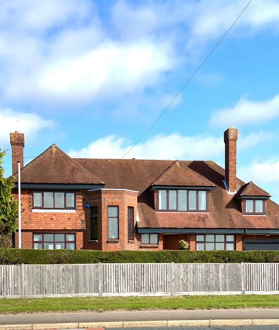 A view of two adjacent residential houses situated in a suburban neighbourhood under a clear blue sky with some light clouds. The house on the left features a brown brick exterior with white-framed windows and a tiled roof with a slight overhang. The house on the right has a combination of brick on the ground floor and white cladding on the upper floor, also with a tiled roof. In front of the houses, a driveway is visible with a silver car parked towards the lower left corner. The foreground shows a small garden with a young shrub and a concrete pathway leading to the entrance doors. Behind the houses, there are tall trees with bare branches and more distant trees with sparse leaves, suggesting the season is late autumn or early winter. The scene is illuminated by natural daylight, highlighting the textures of the brickwork, roofing tiles, and foliage, consistent with a typical property scene that could involve private waste collection or on-site clearance activities by rubbish removal services such as Rubbish Clearance Kingston.