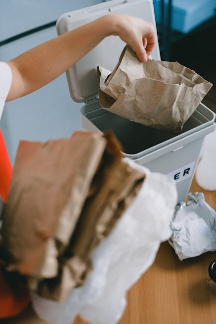 A close-up of a person's hand placing a crumpled brown paper bag into a grey waste bin with the label partially visible, indicating it is used for rubbish disposal. The person is wearing a short-sleeved garment, and the bin is positioned on a wooden surface next to a white wall or appliance. In the foreground, several additional crumpled paper bags are being held, ready for disposal. The scene depicts a typical household or office waste management scenario, illustrating the process of independent rubbish collection. The environment is well-lit, with natural or artificial light highlighting the textures of the paper and the smooth surface of the bin. This imagery correlates with the services provided by Rubbish Clearance Kingston, emphasizing on-site or private waste handling options for domestic or commercial rubbish removal, and aligns with themes of recycling and proper disposal practices in Kingston.
