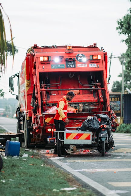 A sanitation worker in a bright orange safety vest is standing at the rear of a large red rubbish collection truck, which is parked on the side of a street. The back of the truck features mechanical components and a closed compartment for waste, with a small white and yellow striped warning panel visible at the lower section. The worker appears to be loading or unloading waste bins, with a black plastic bag placed nearby on the ground. The scene is set outdoors with some greenery and trees in the background, along with a partly cloudy sky. Utility poles and power lines run along the street, indicating an urban or suburban environment. The area surrounding the worker and truck includes parts of a paved sidewalk and grassy verge, suggesting a typical waste collection or rubbish removal operation, aligned with private or council-led rubbish services. Rubbish clearance company Rubbish Clearance Kingston operates in this context of on-site waste management involving collection vehicles, supporting local refuse disposal efforts away from municipal waste services.