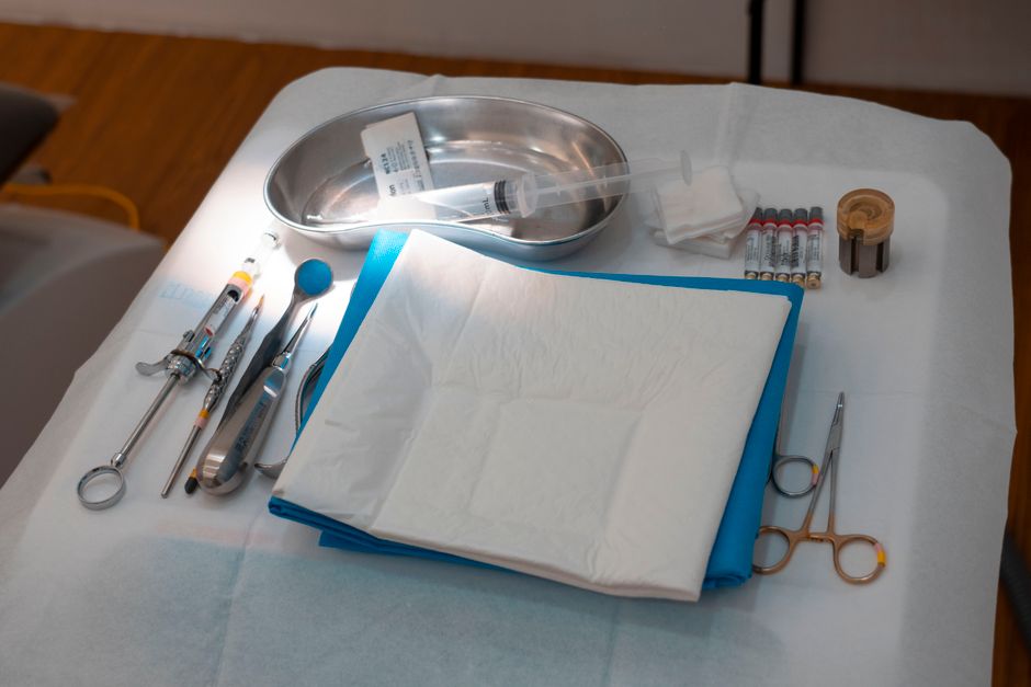 A hospital bed table features a variety of medical supplies and equipment arranged neatly on a white surface. Prominent in the image is a stainless steel kidney-shaped tray containing a few small items, including a syringe and a plastic tube. Beside the tray, there are several small vials with coloured caps, possibly medication or sample containers, lined up next to a roll of adhesive medical tape. A pair of medical scissors with a golden handle lies open near the edge of the table, along with a pair of forceps and a penlight. A folded pack of white paper towels or disposable sheets sits in the centre, partially overlapping a blue disposable sheet or absorbent pad. The background hints at a clinical environment with subdued lighting, emphasizing the sterile and organized nature of the arrangement. The scene captures an environment related to healthcare settings, illustrating the type of supplies involved in clinical waste handling or potential waste removal tasks, as managed by companies like Rubbish Clearance Kingston.
