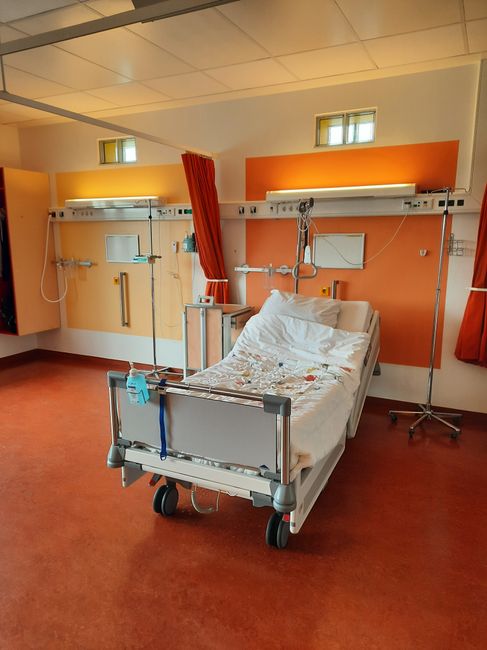 A hospital room featuring a single adjustable hospital bed positioned centrally, with white metal side rails and a mattress covered by a white sheet and some debris or fabric remnants. The bed is on caster wheels and situated on a red-brown floor. Behind the bed, the wall is painted in a warm orange tone, with two small, rectangular windows near the ceiling allowing natural light to enter. Above the bed, a white medical gas and electrical supply panel is affixed to the wall, equipped with various outlets and tubing. To the right of the bed, a vertical IV stand with a hooked arm is present, alongside medical equipment mounted on the wall, including a small monitor and support rails. Red curtains are drawn back on either side, revealing additional clinical fixtures and a neutral ceiling with embedded lighting. The scene suggests a healthcare environment prepared for patient care, and the presence of the hospital bed and medical fittings aligns with the context of clinical waste or rubbish removal, as handled by services such as Rubbish Clearance Kingston.