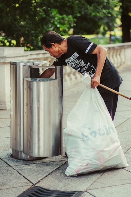 An elderly man with dark hair, dressed in a black T-shirt with white text and dark trousers, is seen leaning forward to dispose of rubbish into a modern, stainless steel public litter bin with a rounded, vertical design positioned on a paved sidewalk. The man holds a large white plastic bag filled with waste in his left hand, while his right hand is lowered toward the opening of the bin. He is using a wooden stick to assist in guiding the rubbish into the container. The background features a low stone balustrade and lush green trees, indicating an outdoor urban environment. The scene is illuminated by natural daylight, and the overall setting suggests a routine litter collection or disposal activity, characteristic of local waste management efforts or private rubbish removal services such as those provided by Rubbish Clearance Kingston. The image underscores the importance of maintaining cleanliness in public spaces through responsible rubbish disposal and the role of independent or commercial waste collection solutions.