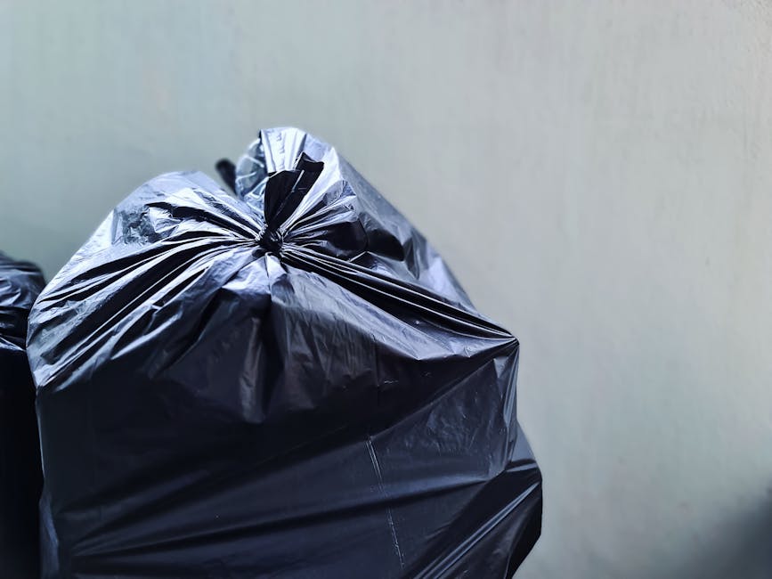 A black wheeled rubbish bin with the label 'St. John’s' on the side, positioned on a paved sidewalk beside a street at night. The bin is open, revealing stacked, partially crushed cardboard boxes and assorted waste materials inside. The street is illuminated by warm streetlights, casting soft shadows on the pavement and nearby curb. Behind the bin, there are trees and dark hedges lining the edge of the sidewalk, with some foliage partially visible under the street lighting. In the background, the street extends into the distance with additional streetlights and parked vehicles faintly visible, giving an urban setting that suggests this scene relates to private waste collection or rubbish removal services such as those provided by Rubbish Clearance Kingston. The overall scene emphasizes the importance of proper waste handling and the use of designated disposal bins for rubbish collection outside of regular municipal waste services, aligning with the company's focus on independent rubbish clearance and waste management solutions.