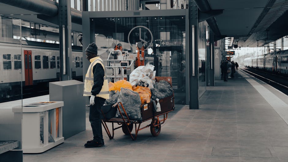 A worker standing on a modern outdoor train station platform, dressed in dark clothing, a high-visibility vest, and a grey beanie, is positioned beside a red flatbed trolley overloaded with various waste bags and loose rubbish, including black, grey, and bright orange plastic bags filled with discarded materials. The trolley is situated close to a glass shelter, partially reflecting the surrounding area. In the background, there are train tracks with passenger trains parked alongside, and a few individuals are seen further along the platform, some standing and others walking. The scene is illuminated by natural daylight, with an urban environment encompassing sleek, contemporary architecture, and the platform's smooth grey paving extends into the distance. This setting emphasizes the importance of proper rubbish handling and disposal, which companies like Rubbish Clearance Kingston facilitate through alternative waste collection services, ensuring efficient removal of waste from public areas such as train stations or urban transport hubs.