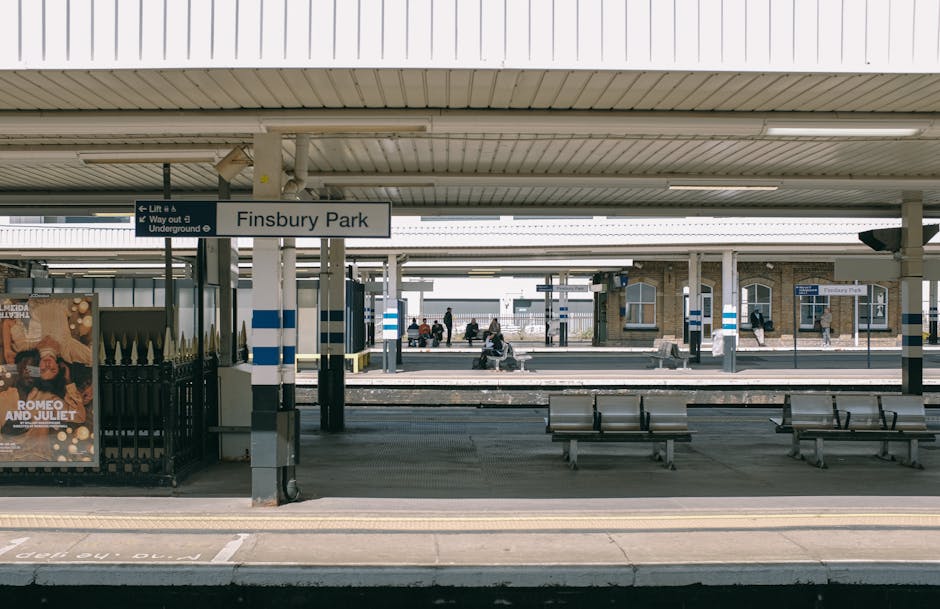 The image depicts a railway station platform under an extensive metal and glass canopy structure. On the left side, there is a historic brick building with multiple windows and a small staircase leading down from the platform. Several passengers are visible standing or sitting along the platform, waiting for the train, while some are engaged in conversation. The railway tracks run horizontally across the foreground, with gravel ballast surrounding the rails. The platform is lined with tactile paving in a yellowish tone to assist visually impaired travelers, and the surface appears to be made of concrete slabs with sections painted in yellow for safety and guidance. Overhead, metal beams and a glass roof provide shelter, allowing natural light to filter through, creating a bright but shaded environment. At the far end of the platform, signage indicates directions for exits and platforms. The overall scene suggests a well-maintained, functional transport hub, where private or independent transport options, such as rubbish clearance services like Rubbish Clearance Kingston, might coordinate to serve passengers traveling through the station area.