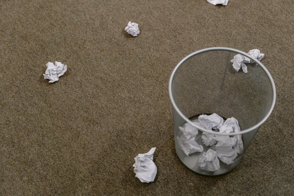 A clear, cylindrical waste bin made of lightweight plastic with a smooth, semi-transparent surface sits on a brown carpeted floor. The bin contains several crumpled sheets of white paper, with some pressed against the inner walls and others piled loosely at the bottom. Surrounding the bin, multiple additional crumpled paper balls are scattered across the carpet, varying in size and tightly or loosely crumpled, creating a disorganized appearance. The environment appears to be an indoor space with even, soft lighting, highlighting the textures of the paper and the carpet. The scene suggests a setting where informal or temporary rubbish collection occurs, possibly aligned with private waste handling services such as those provided by Rubbish Clearance Kingston, emphasizing a simple, on-site clearing or disposal process within a domestic or office environment. The overall scene emphasizes practical waste management with straightforward, accessible rubbish removal in a non-industrial setting.
