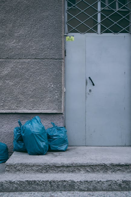 A large pile of mixed household waste and rubbish sits on the ground beside a low stone wall, with black plastic bags, cardboard boxes, and discarded items visible. A yellow plastic container is partially buried under the debris, while an old, worn car seat with light beige fabric upholstery leans against the rubbish and the stone wall. The waste appears to contain various materials, including plastic, paper, and fabric, indicative of domestic refuse awaiting collection or removal. In the background, there is a metal fence, some greenery, and a structure with a curved roof, all under a clear blue sky with a few wispy clouds. The scene is outdoors, possibly on a driveway or service area, highlighting a typical scenario where independent rubbish clearance services might be employed for bulky or tight-access waste removal, as provided by Rubbish Clearance Kingston. The environment demonstrates an unmanaged disposal site that requires professional intervention to ensure proper clearance, especially where access is limited or waste volume is significant.