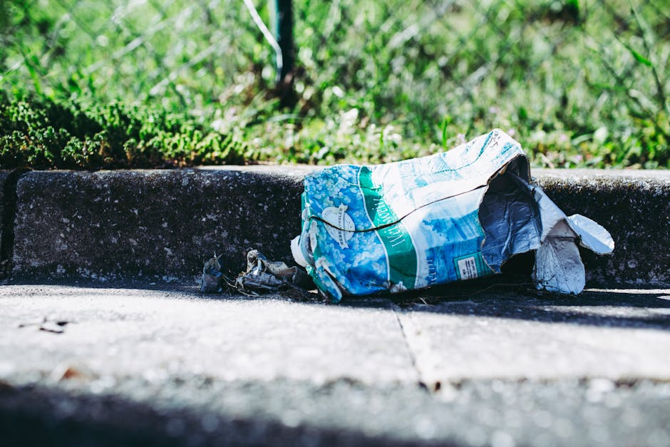 A black wheeled rubbish bin labeled 'St. John's' is positioned on a sidewalk at night, with the lid open and overflowing with various waste items, including brown cardboard boxes, white paper, and plastic bags. The bin is placed next to a curb along a quiet urban street illuminated by streetlights, with trees and shrubbery lining the pavement on the right. In the background, there are faint outlines of parked cars and distant street lighting, creating a dimly lit atmosphere typical of late evening or night. The scene reflects a context where waste has been temporarily stored for collection, possibly indicating the need for waste removal service through private disposal methods, such as those provided by Rubbish Clearance Kingston, rather than council-managed rubbish collection. The ambient light and urban setting emphasize the importance of proper rubbish management and highlight the types of waste that may require professional waste clearance services to maintain cleanliness and compliance with local regulations.